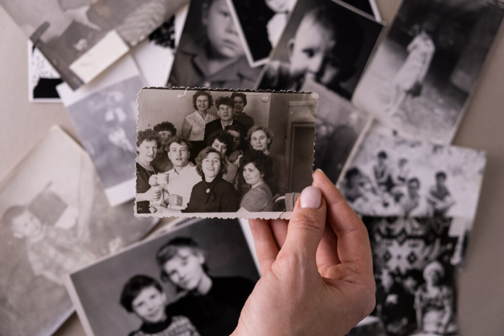 A hand delicately holds a vintage black and white photograph depicting a group of young adults and adults, surrounded by other old photos, evoking a sense of nostalgia and remembrance. The monochrome palette emphasizes the historical quality of the images.
