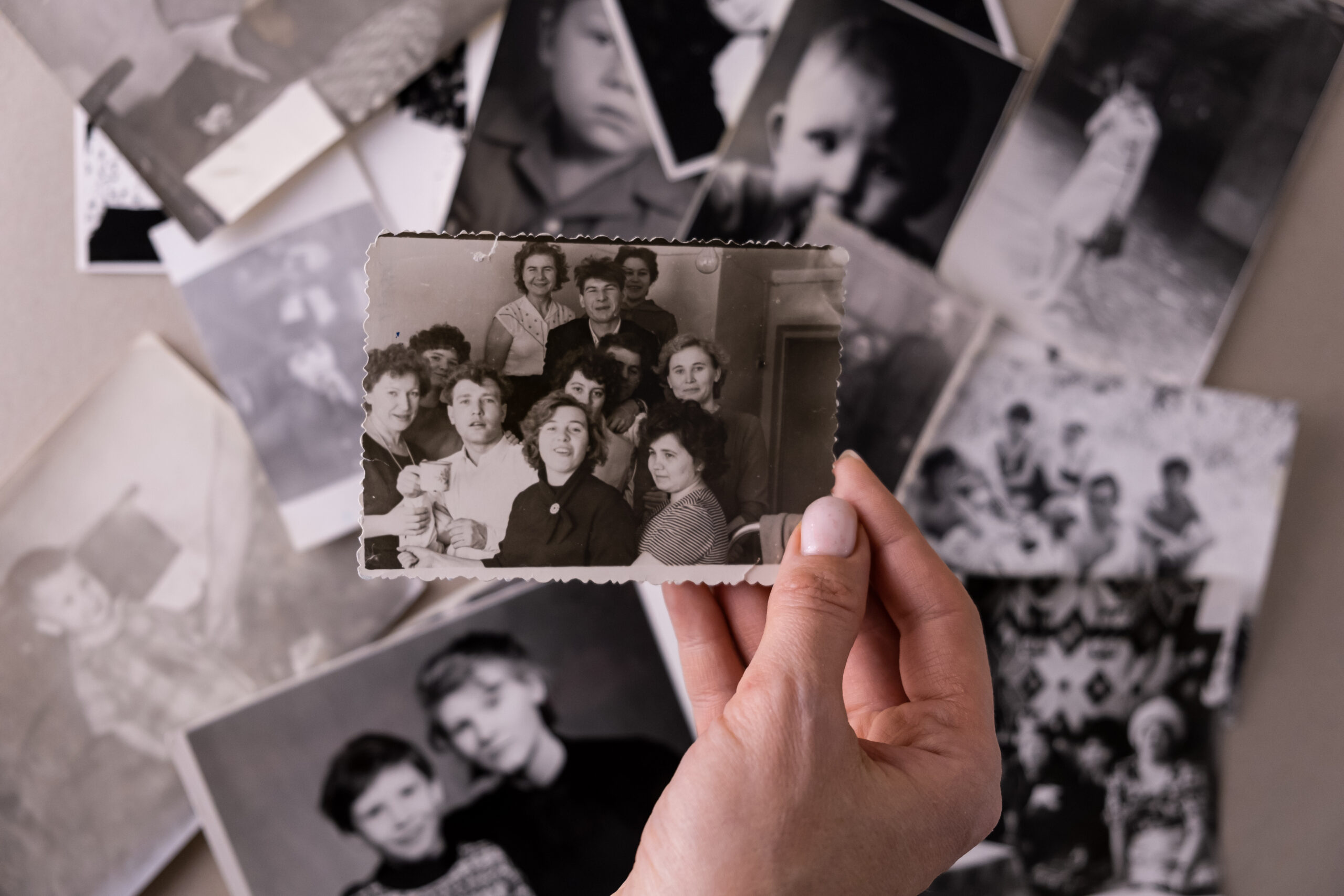 A hand delicately holds a vintage black and white photograph depicting a group of young adults and adults, surrounded by other old photos, evoking a sense of nostalgia and remembrance. The monochrome palette emphasizes the historical quality of the images.
