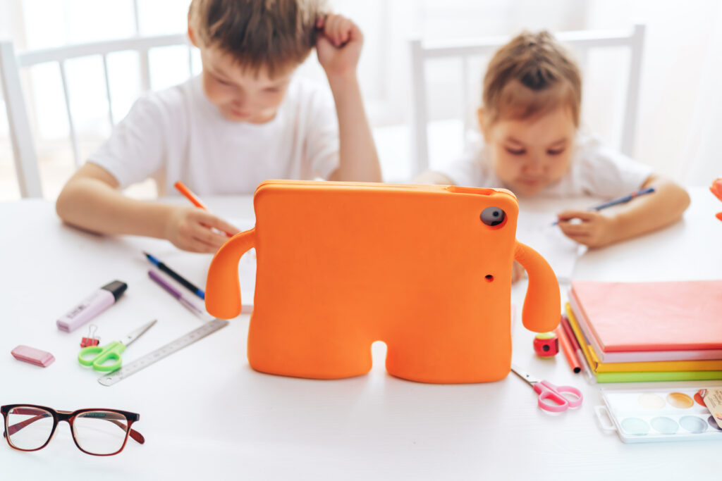 Two children with light hair and white shirts sit at a table, concentrating on drawing or writing on paper with colorful pens. In the foreground is an orange tablet case with arm-like features. Various art supplies, including pens, pencils, scissors, watercolor paints, and notebooks, are scattered across the white table.